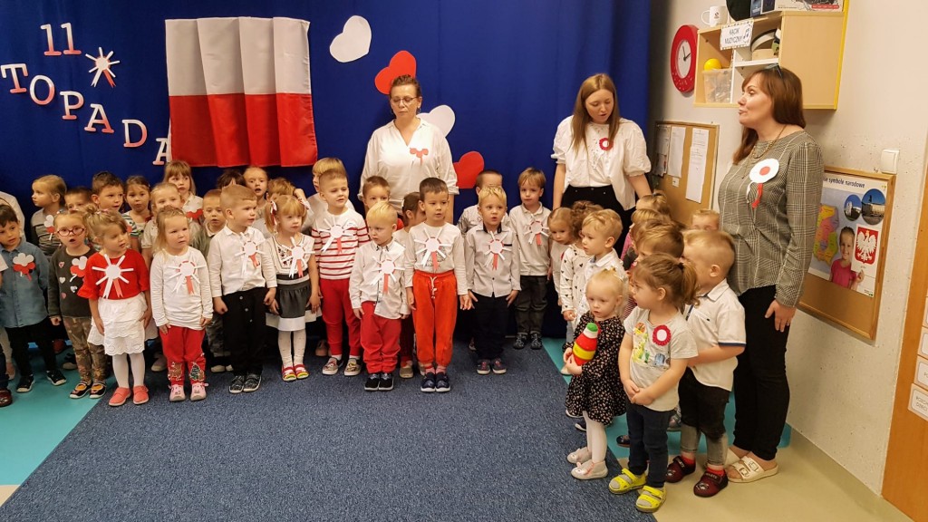 Dzieci na tle bialo czerwonej flagi spiewaja hymn polski