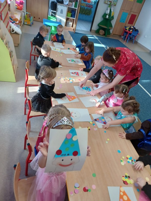Dzieci siedza przy stolikach i przyklejaja kolorowe kropki na czapeczki