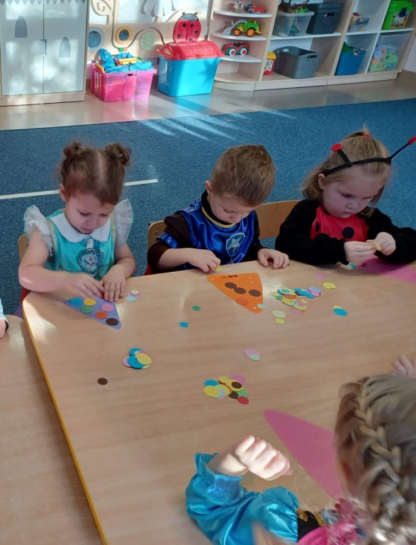 Dzieci siedza przy stoliku i przyklejaja kolorowe kropki na czapeczki od pajaca
