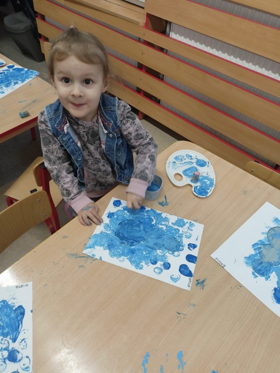 Dziewczynka siedzi przy stoliku i maluje niebieska farba
