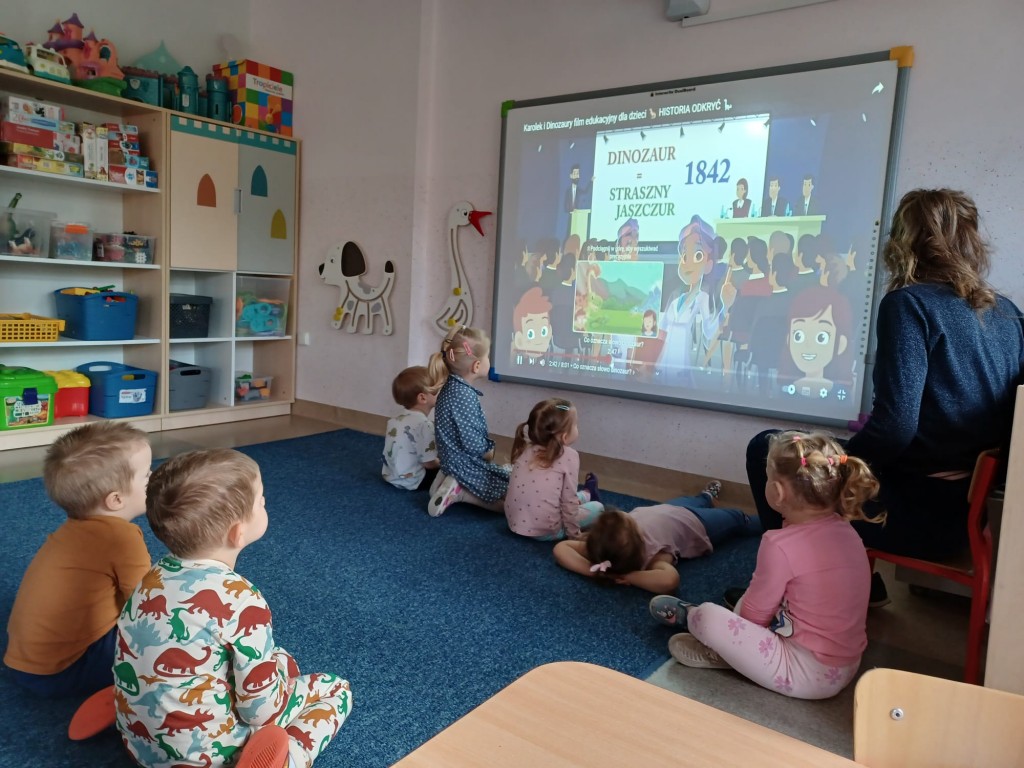 dzieci siedza na dywanie i ogladaja film edukacyjny o dinozaurach
